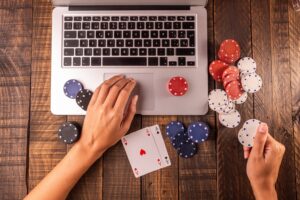 Ein Laptop steht auf einem hölzernen Tisch mit Pokerchips, Spielkarten und zwei Händen. 