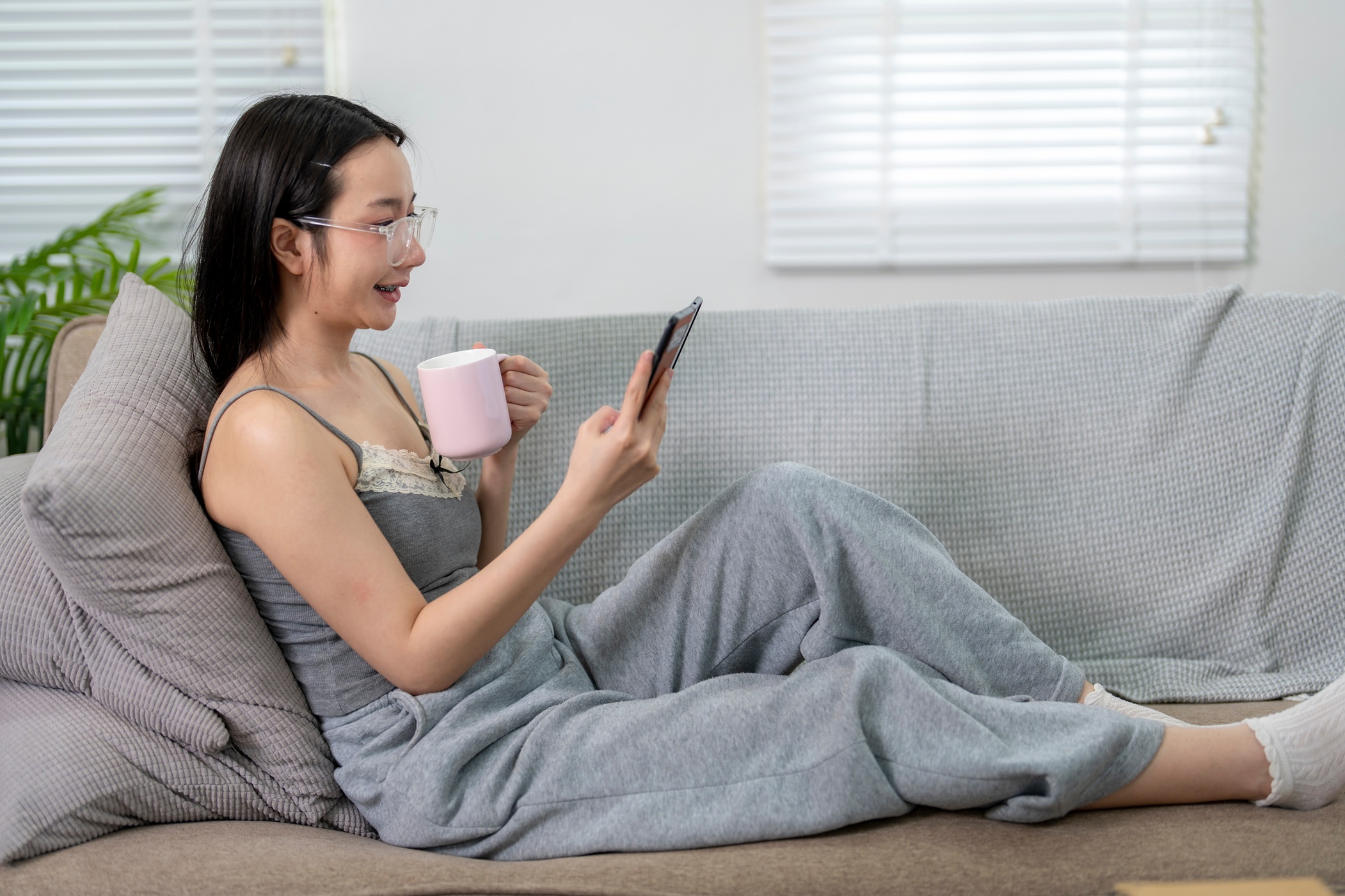 Junge Frau in Jogginghose sitzt mit Kaffeetasse und Smartphone auf dem Sofa.